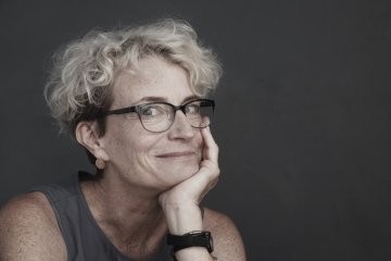 Photo of Ashton Applewhite, a white woman with short, curly blond hair and glasses. She is holding her head in her left hand and looking sideways at the camera. Background is gray. 