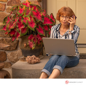 Photo of a white woman sitting on the front step of a house with a laptop on her lap and a cell phone in her left hand, held to her ear. She has red hair, glasses, and is wearing a striped blue shirt with jeans rolled up at the ankles. A large plant with red and green leaves sits to her right.