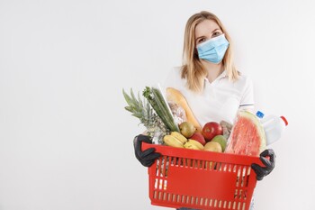 Image of a white woman with blond hair and a disposable blue face mask holding a shopping basket filled with groceries including fresh fruit and vegetables and meat and dairy products. 