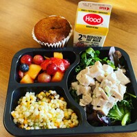Photo of a meal. At top, a muffin sits next to a carton of 2% Hood milk. Below, a food tray with three sections. Top left shows mixed, cut fruit. Below, couscous salad. Right, larger green salad with chicken on top.