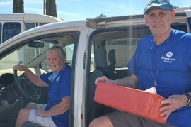 Photo shows a woman with light skin and white hair pulled back sitting in the driver's seat of a minivan with the door open. An older man with light skin and a blue hat with a  stands in the side door area of the van, stepping out and holding a box. Both wear blue AmeriCorps Seniors t-shirts