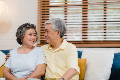 photo of an older woman with gray hair and a blue t-shirt sitting with an older man who has gray hair and a yellow button down shirt. They are sitting together on a couch, with a window behind them, wooden blinds open, looking at each other. The man has his arm around the window.