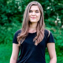 Photo of a woman with light brown hair that goes just past her shoulders. She is wearing a black t-shirt and smiles at the camera. Background shows trees, grass, and bushes.