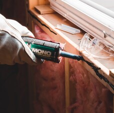 Photo shows a leather-gloved hand applying caulk to a windowsill, where the wall below is still being insulated.